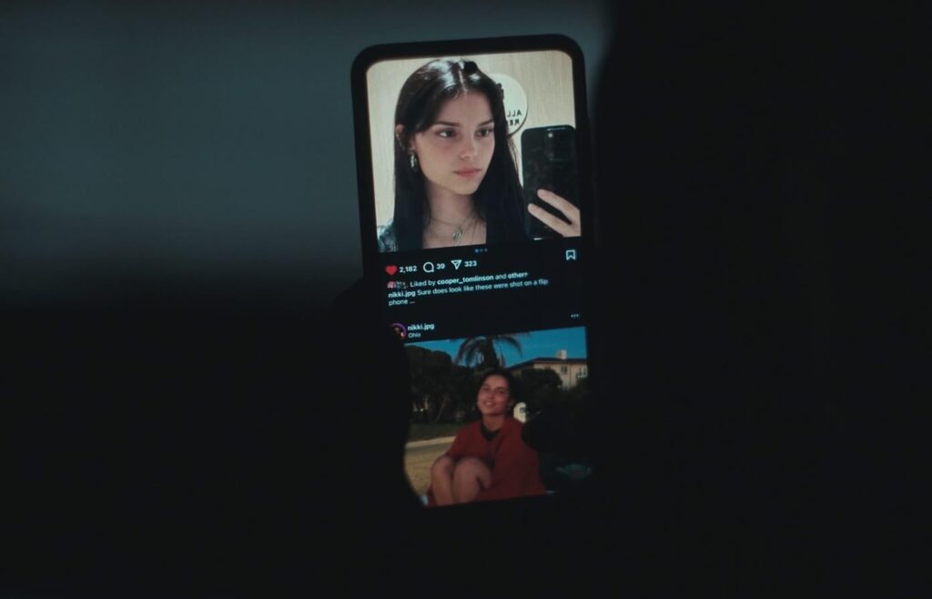 Eine Nahaufnahme eines Smartphones, das in dunkler Umgebung gehalten wird und einen Social-Media-Feed zeigt. Auf dem Display ist oben das Spiegel-Selfie einer jungen Frau mit dunklen Haaren zu sehen, darunter ein weiteres Foto von ihr im roten Oberteil im Freien. Das aufleuchtende Display im schattigen Umfeld verdeutlicht das Stalken und die digitale Fixierung auf die abgebildete Person. | FYP Filme