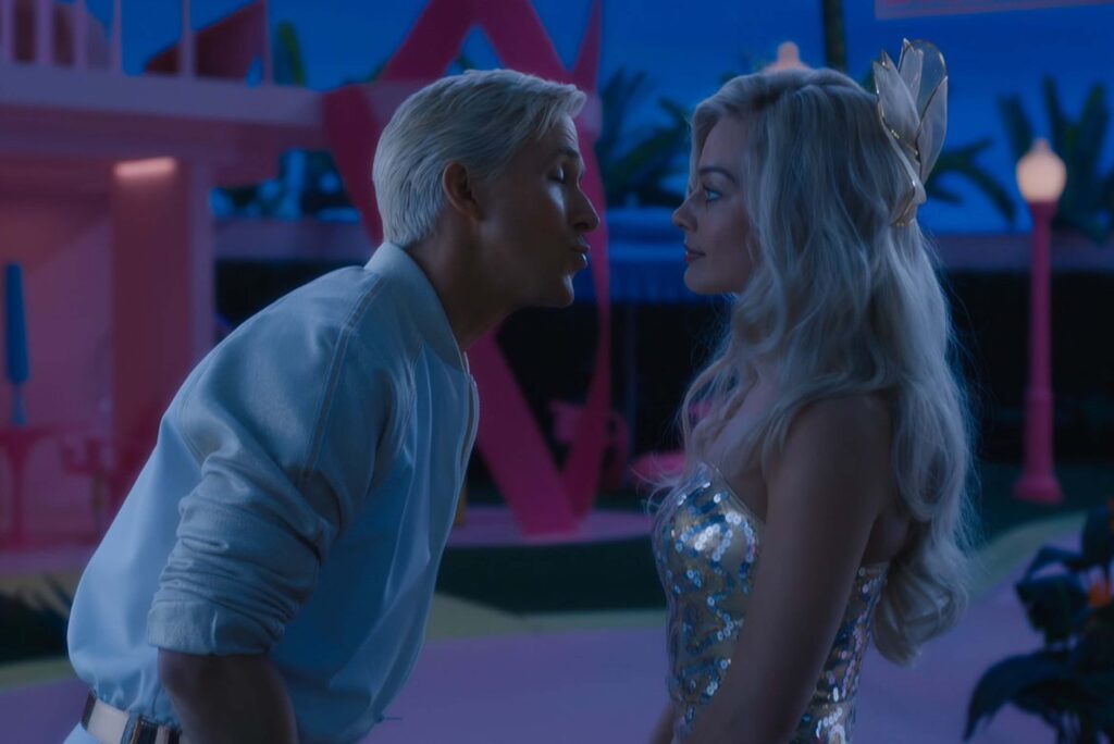 Ryan Gosling als Ken und Margot Robbie als Barbie stehen eng nebeneinander in einer farbenfrohen, stilisierten Umgebung. Ken trägt eine weiße Jacke und hat kurzes blondes Haar, während Barbie ein glitzerndes, schulterfreies Kleid und langes, welliges blondes Haar trägt. Hinter ihnen leuchten pinke und blaue Farbtöne, Palmen und verspielte Architektur, was der Szene einen traumhaften, leicht futuristischen Barbie‑Look verleiht. | Media Monday #790