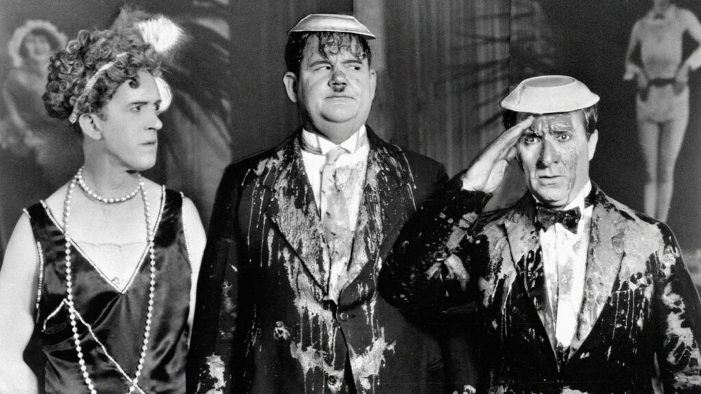 Das ist meine Frau (1929), in der Dick und Doof nebeneinander stehen, beide mit Melonenhüten und von oben bis unten mit Essen oder Flüssigkeit bespritzt. Neben ihnen steht Vivian Oakland als Hardys Filmfrau, ebenfalls völlig beschmutzt und mit Federkopfschmuck. Dick salutiert ernst, während Doof verwirrt wirkt; im Hintergrund sind unscharfe Tänzerinnen zu sehen. Die Darstellung zeigt den typischen Slapstick‑Humor des Duos Dick und Doof.
