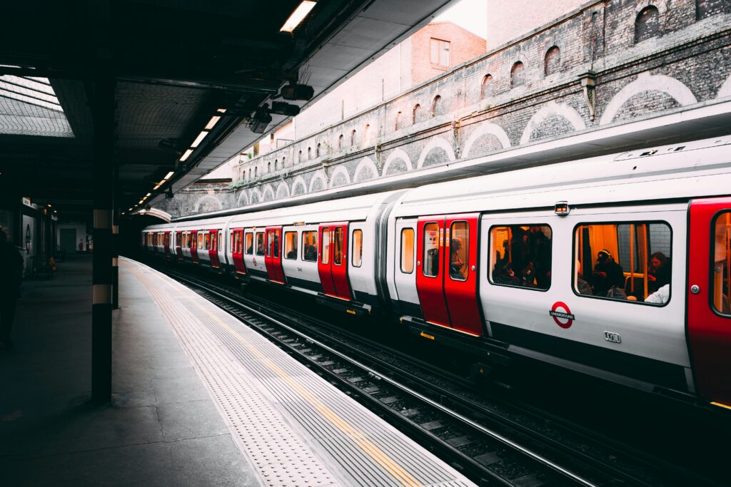 Ein Zug der London Underground steht an einem Bahnsteig. Die silbernen Wagen mit roten Türen tragen das typische London‑Underground‑Logo. Der Bahnsteig ist teilweise überdacht, mit Beleuchtung und einem gelben Blindenleitstreifen am Rand. Im Hintergrund sind gemauerte Bögen und darüberliegende Gebäude zu sehen. | Media Monday #788