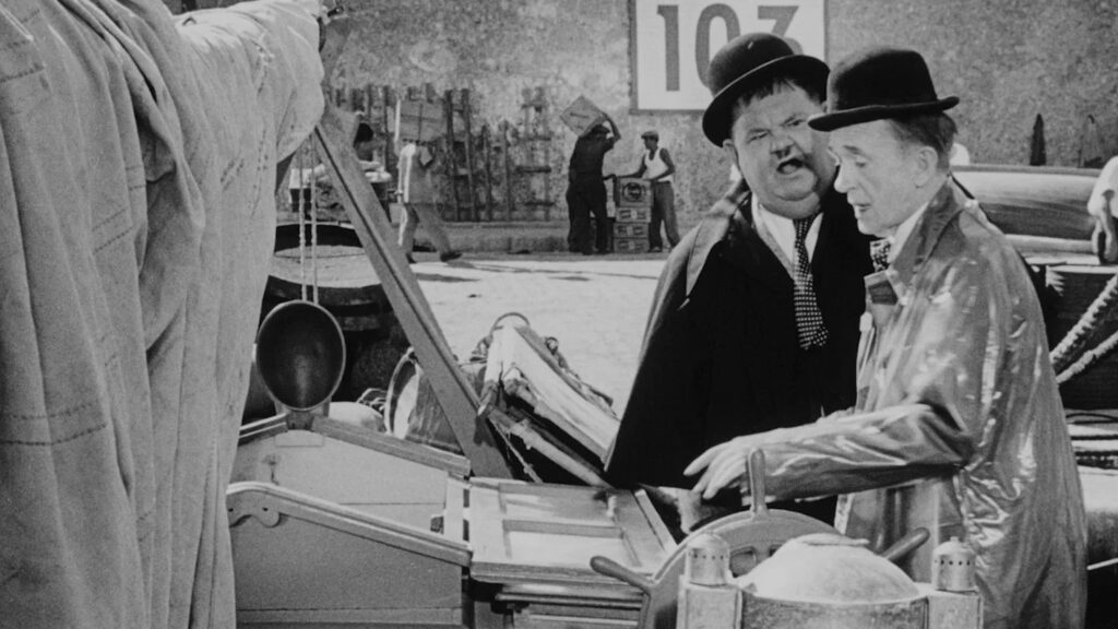 Szene aus Dick und Doof erben eine Insel, in der Laurel und Hardy mit ihren typischen Melonenhüten neben einem mechanischen Gerät stehen. Laurel reagiert gestenreich und überrascht, während Hardy aufmerksam auf die Apparatur blickt. Im Hintergrund tragen Arbeiter Kisten vor einer Wand mit der großen Zahl „103“. Die Darstellung zeigt die klassische Slapstick‑Dynamik des Duos, gespielt von Laurel und Hardy.