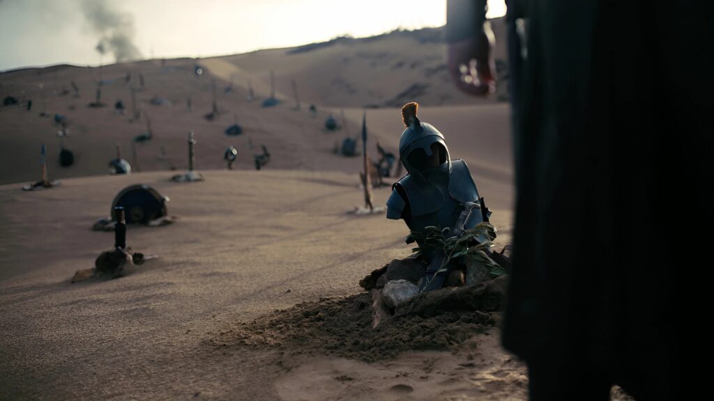 Ein einzelner Helm und verstreute Waffen ragen aus den Dünen eines verlassenen Schlachtfeldes, während eine Gestalt schweigend vorbeigeht. Das eindringliche Motiv symbolisiert die Vergänglichkeit von Ruhm und die bleibenden Spuren des Krieges.  | Die Odyssee