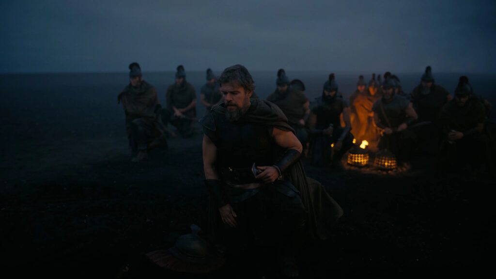 Odysseus (Matt Damon) kniet erschöpft auf einem dunklen Schlachtfeld, während seine Männer schweigend hinter ihm warten. Die Szene vermittelt die Last des Krieges und den Beginn seiner langen Heimreise. | Die Odyssee