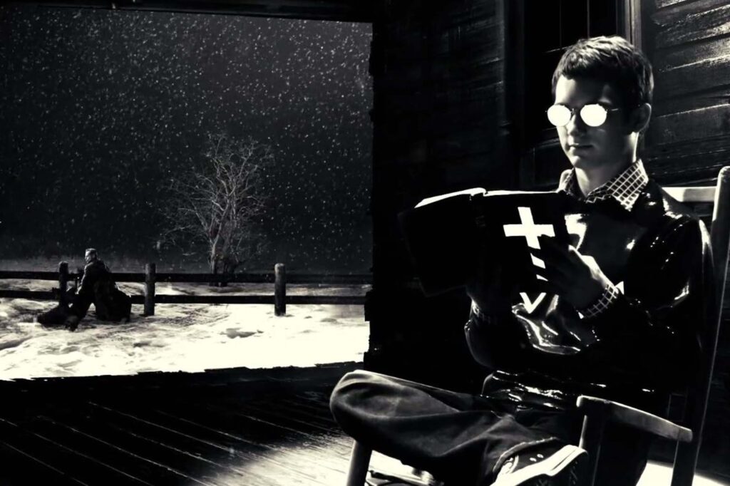 Kevin (Elijah Wood) sitzt in einer dunklen Holzhütte auf einem Stuhl und liest ein Buch mit einem weißen Kreuz auf dem Einband, während draußen dichter Schnee fällt. Durch die offene Tür sieht man Hartigan (Bruce Willis), der im verschneiten Hof nahe eines Zauns kniet. Die Szene ist stark kontrastreich und wirkt wie ein monochromes Noir‑Bild aus dem Film Sin City. | Media Monday #792