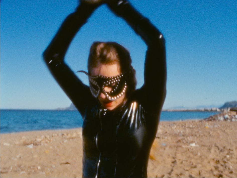 Lilith Stangenberg steht maskiert in einem glänzenden schwarzen Outfit am Strand, die Arme über den Kopf gehoben; hinter ihr liegen Meer und ferne Berge unter klarem Himmel – Szene passend zu ihrer Rolle als Charlotte im Film Für immer 16.