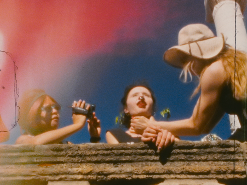 Für immer 16: Charlotte (Lilith Stangenberg), Helena (Mücke) und Oskar (Chang Wang) lehnen hinter einer niedrigen Steinmauer im Freien; Oskar filmt mit einer kleinen Kamera wie Charlotte, Helena am Hals packt während der Himmel klar und hell über ihnen steht.
