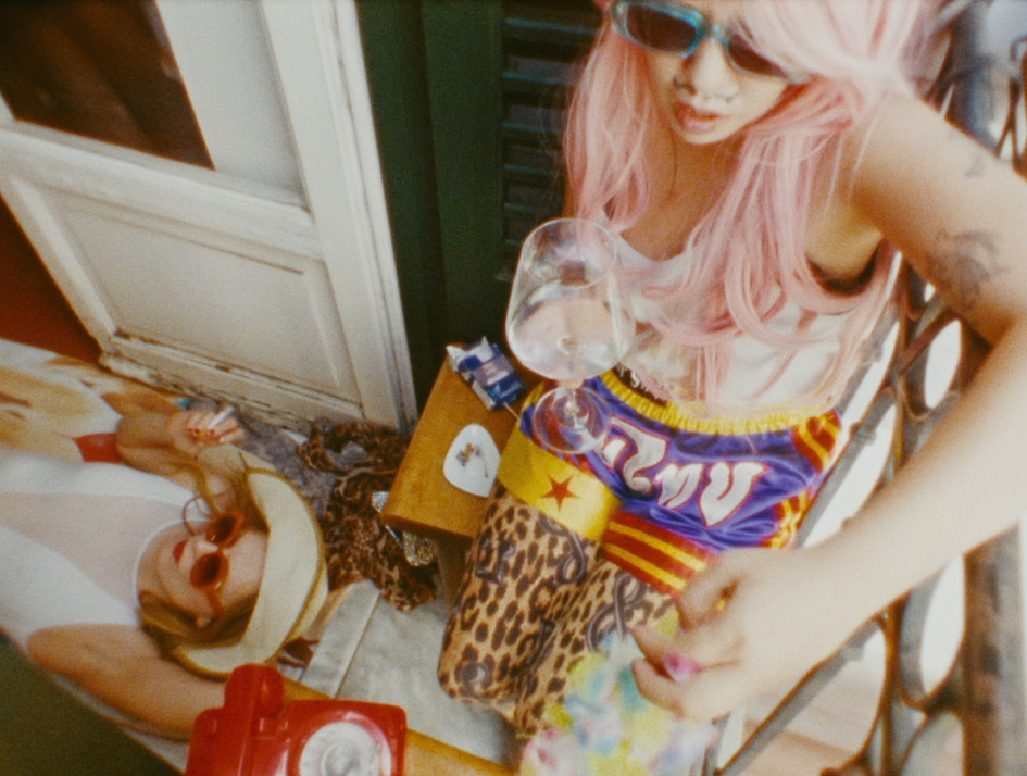 Oskar (Chang Wang) sitzt am Fenster im bunten Outfit, Sonnenbrille und einem Glas Weißwein in der Hand, während Charlotte (Lilith Stangenberg) unten am Boden liegt, mit Sonnenhut, Sonnenbrille und Zigarette. Die Szene ist aus dem Film "Für immer 16" und wirkt ausgelassen