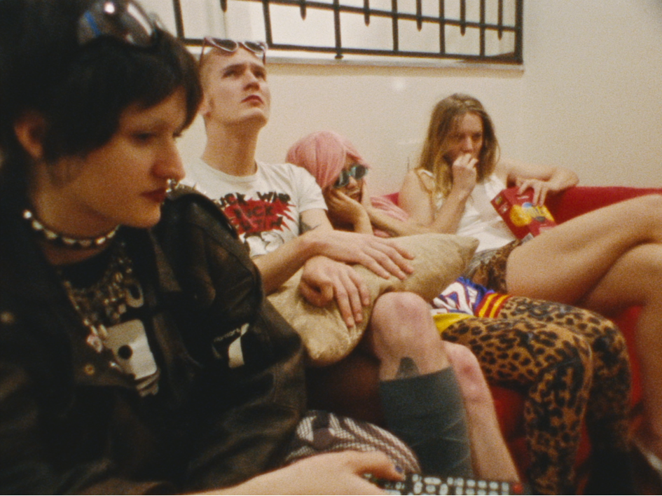 Oskar (Chang Wang), Charlotte (Lilith Stangenberg), Helena (Mücke) und Rudolph (Twiggy Glouglou) sitzen eng beieinander auf einem roten Sofa, ein chaotisch‑intimer Moment aus dem Film Für immer 16: Oskar trägt wie immer Sonnenbrille und pinke Perücke. Helena ihre Lederjacke und Charlotte isst Chips. Klassische Szene aus "Für immer 16"