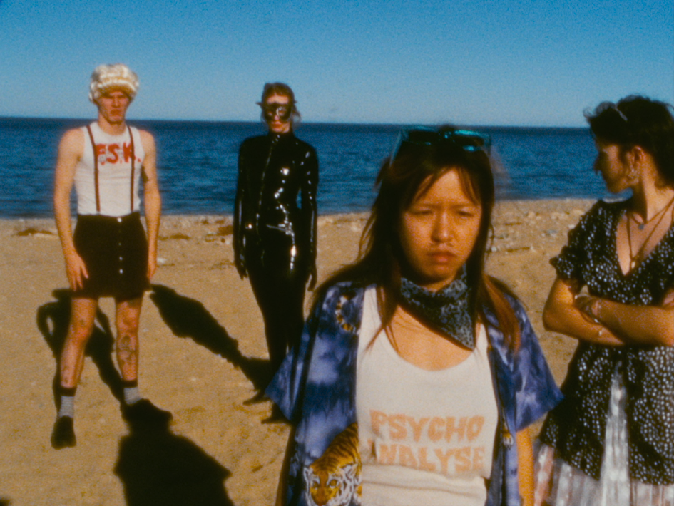 Rudolph (Twiggy Glouglou) steht ganz links am Strand in weißem Tanktop, schwarzer kurzer Hose mit Hosenträgern und weißer Perücke; daneben Oskar (Chang Wang) in blau gemustertem Hemd, Sonnenbrille und „Psycho Analyse“-Shirt, dahinter Charlotte (Lilith Stangenberg) im schwarzen Ganzkörperoutfit, rechts daneben Helena (Mücke) in dunklem Kleid mit verschränkten Armen. Die vier posieren gemeinsam vor Meer und Himmel