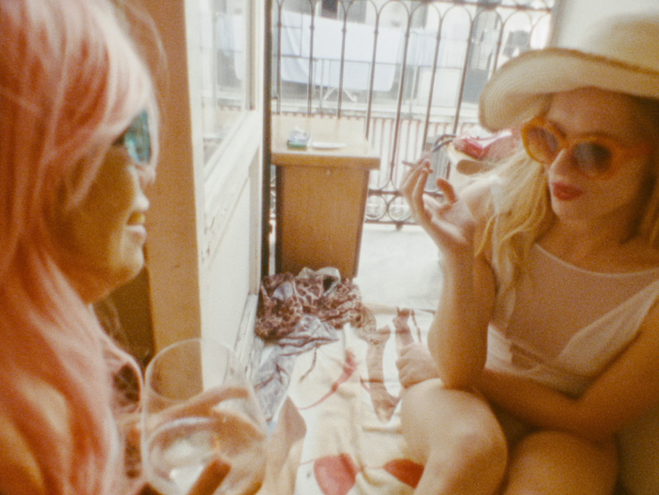 Oskar (Chang Wang) mit pinken Haaren und blauen Sonnenbrillen sitzt links neben Charlotte (Lilith Stangenberg), die einen großen Sonnenhut, orangefarbene Brille und eine Zigarette trägt; beide sitzen entspannt auf einer Decke in einem kleinen Innenraum, während hinter ihnen ein Tisch und Kleidung zu sehen sind – eine ruhige Momentaufnahme aus dem Film Für immer 16.
