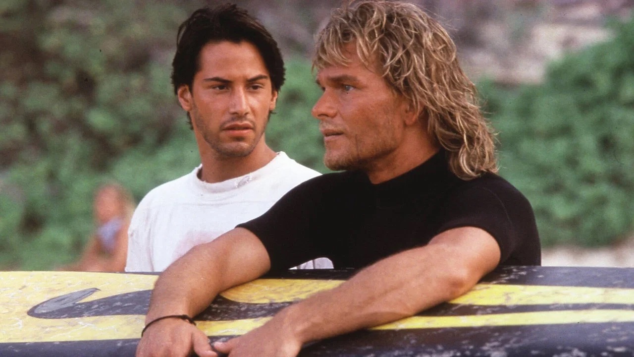 Keanu Reeves als Johnny Utah und Patrick Swayze als Bodhi stehen nebeneinander im Grünen und lehnen an einem gelb‑schwarzen Surfbrett; beide wirken konzentriert und im Gespräch, Szene aus dem Film Gefährliche Brandung. | Top 5 Filme mit exotischen Schauplätzen