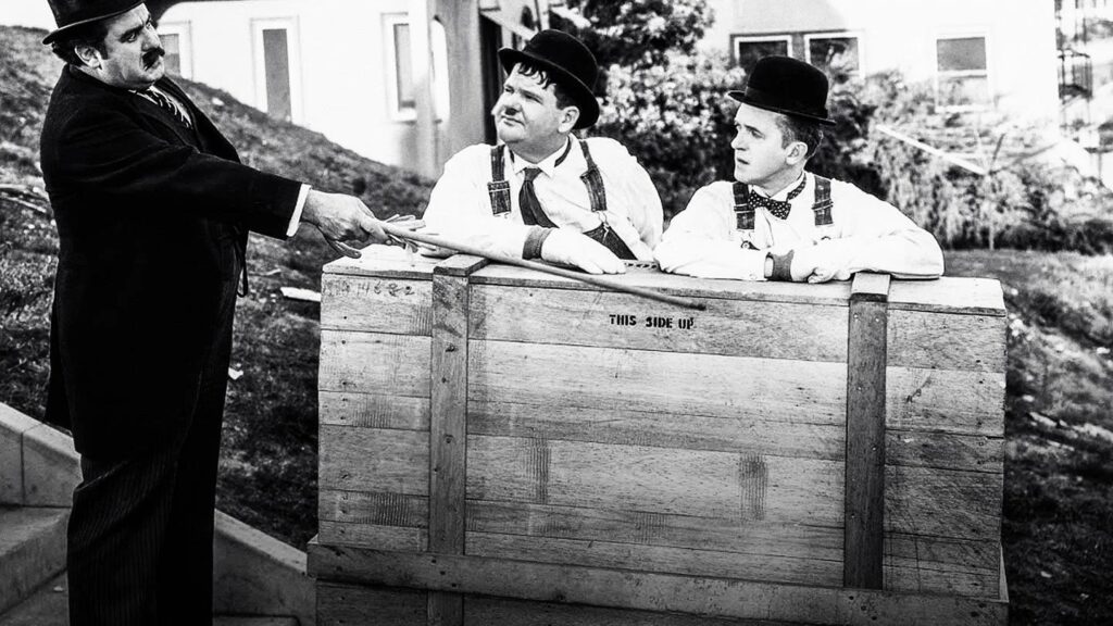 Szene aus Laurel und Hardy: Der zermürbende Klaviertransport mit Laurel und Hardy, die in einer großen Holzkiste sitzen, während ein Mann in dunklem Anzug und Hut mit einem langen Holzbrett auf sie zeigt. Die Kiste trägt die Aufschrift „This Side Up“, im Hintergrund stehen ein Gebäude und eine grasbewachsene Böschung.