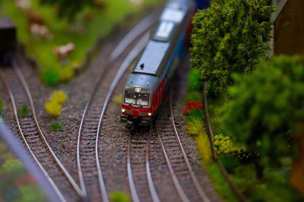 Ein detailreiches Modellbahn‑Diorama mit einer kleinen rot‑grauen Lokomotive, die auf einer gebogenen Schiene durch eine naturgetreu gestaltete Landschaft aus grünen Bäumen, Sträuchern, Grasbüscheln und roten Blumen fährt; im Hintergrund weitere Gleise und kleine Modelltiere wie Kühe. | Media Monday #791