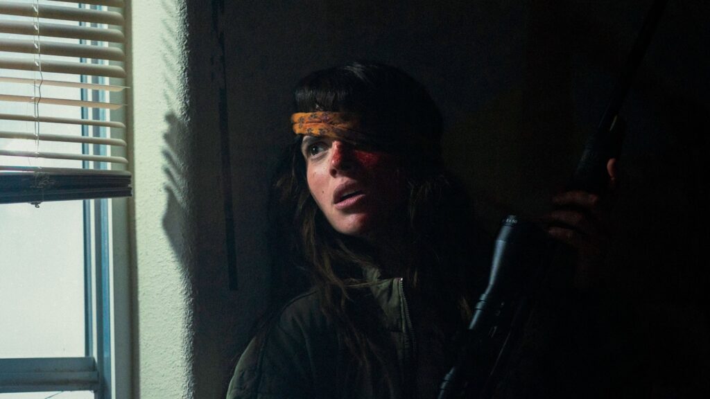 Celeste späht mit Waffe im Anschlag durch ein Fenster raus; ein Kopftuch bedeckt ihr verletztes Auge — Onslaught | 2026 © Plaion Pictures | A24