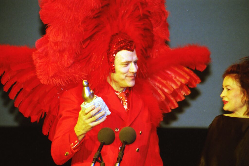 Rosa von Praunheim steht auf einer Bühne beim Teddy Award 1999 im Haus der Kulturen der Welt in Berlin. Er trägt ein auffälliges rotes Kostüm mit großen Federflügeln und hält eine goldene Preisfigur in der Hand. Neben ihm steht eine weitere Person in dunkler Kleidung und blickt zu ihm hinüber. Beide sind vor einem dunklen Hintergrund zu sehen. | Media Monday #788