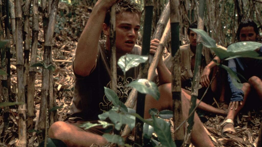 Richard Fischer (Leonardo DiCaprio), Étienne (Guillaume Canet) und Françoise (Virginie Ledoyen) hocken im dichten Dschungel zwischen hohen Bambuspflanzen und verstecken sich angespannt vor bewaffneten Drogenkriminellen; eine Szene aus The Beach, geprägt von Angst und unmittelbarer Gefahr. | Top 5 Filme mit exotischen Schauplätzen