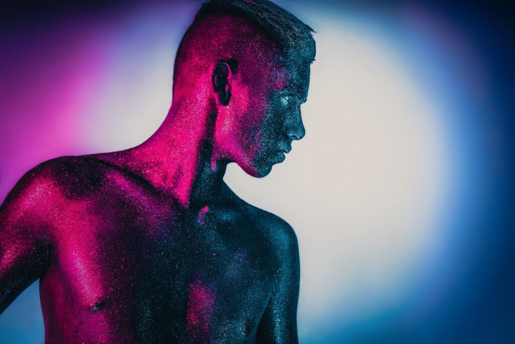 Eine Person, deren Oberkörper und Gesicht vollständig mit glitzernder, metallischer Farbe bedeckt sind, steht seitlich im Bild. Intensives pinkes und blaues Licht fällt auf die Haut und erzeugt starke Kontraste. Der Hintergrund verläuft weich von Pink zu Blau und verstärkt den futuristischen, künstlerischen Look der Aufnahme. | Media Monday #790
