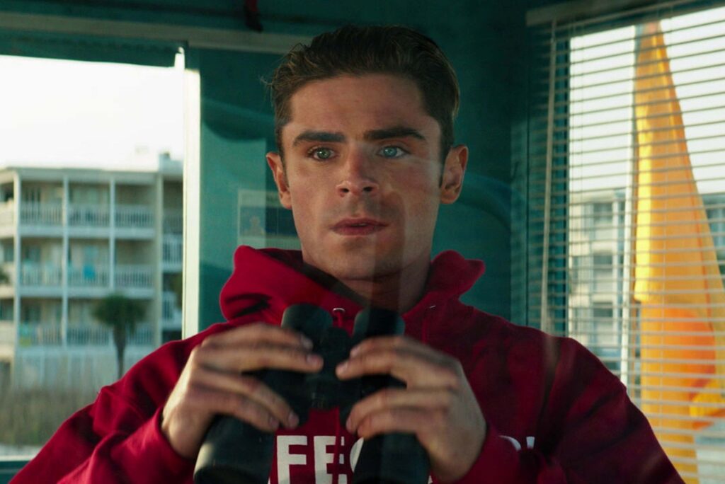 Zac Efron als Brody steht in einem Wachraum am Fenster, trägt einen roten Hoodie und hält ein Fernglas in den Händen. Durch die Jalousien fällt Tageslicht auf ihn, während draußen mehrstöckige Gebäude und Palmen zu sehen sind, was die typische Baywatch Atmosphäre eines Küstenorts vermittelt. | Media Monday #790
