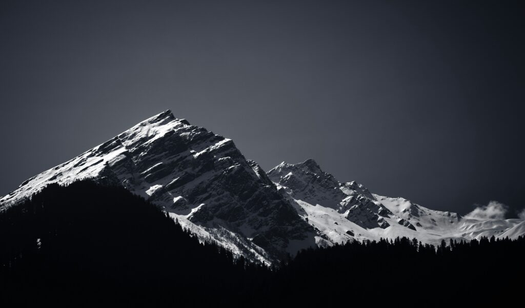 Dunkle, dichte Baumreihe im Vordergrund, dahinter steile, schneebedeckte Bergspitzen unter einem klaren Himmel. Die starke Licht‑Schatten‑Kontrastierung betont die rauen Felsstrukturen und die dramatische alpine Landschaft. | Media Monday #793