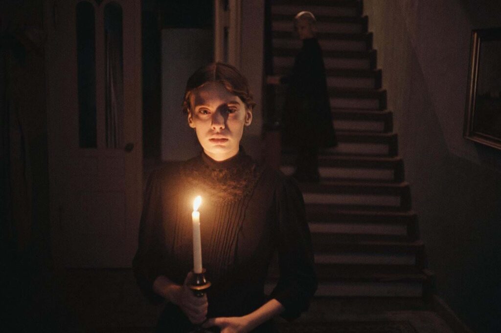 Das Bild zeigt eine Szene aus dem Film "In die Sonne schauen". Ein Mädchen steht mit einer Kerze in einem dunklen Flur. Im Hintergrund führt eine Treppe nach oben. Dort steh ein junges Mädchen. | Themenübersicht Oktober 2026