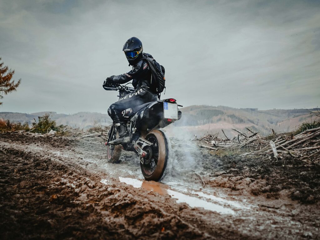 Motorradfahrende Person in schwarzer Schutzkleidung fährt durch eine matschige Offroad-Strecke. Das Hinterrad spritzt Wasser und Schlamm auf, während im Hintergrund kahle Hügel, Baumstümpfe und ein bewölkter Himmel zu sehen sind. Die Aufnahme vermittelt Bewegung und Abenteuer in einer rauen Naturlandschaft. | Media Monday #796