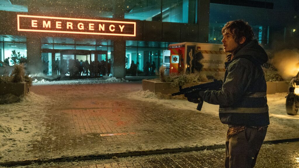 Das Bild zeigt Bryan (Austin Abrams) aus dem Film RESIDENT EVIL. Er hält ein Maschinengewehr vor sich. Im Hintergrund sieht man ein Gebäude mit einem Leuchtschild auf dem EMERGENCY steht. Darunter sind mehrere Menschen hinter einer Glasfront zu sehen