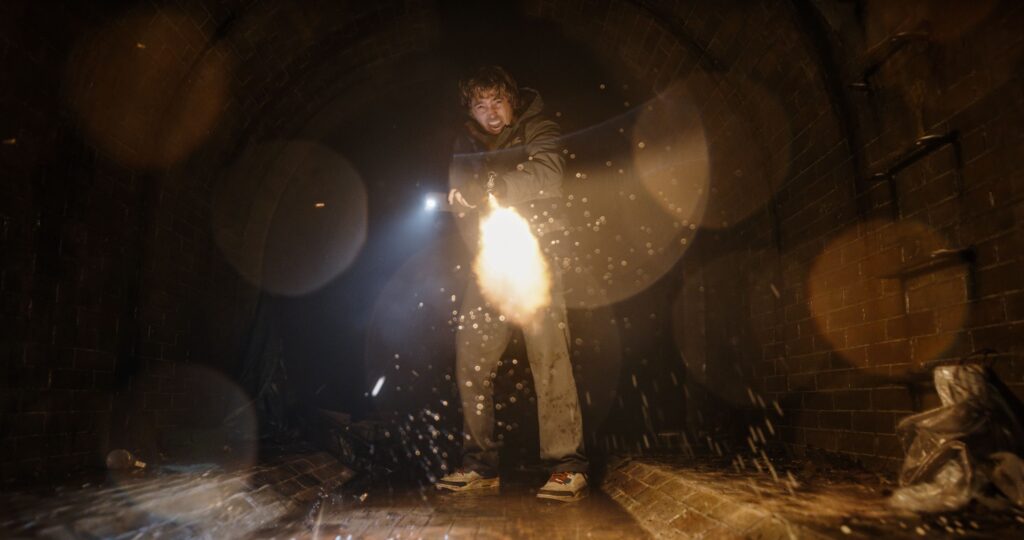 Das Bild zeigt Bryan (Austin Abrams) in einem gekachelten Gang in Dunkeln. Er feuert ein Maschinengewehr ab