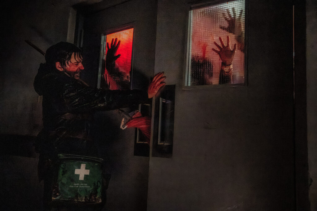 Das Bild zeigt Bryan (Austin Abrams) mit blutverschmierten Gesicht und einer Rescue Tasche umgeschlungen. Er stemmt eine Doppeltür zu, die zwei Scheiben hat. An den Scheiben sind mehrere blutende Hände zu sehen