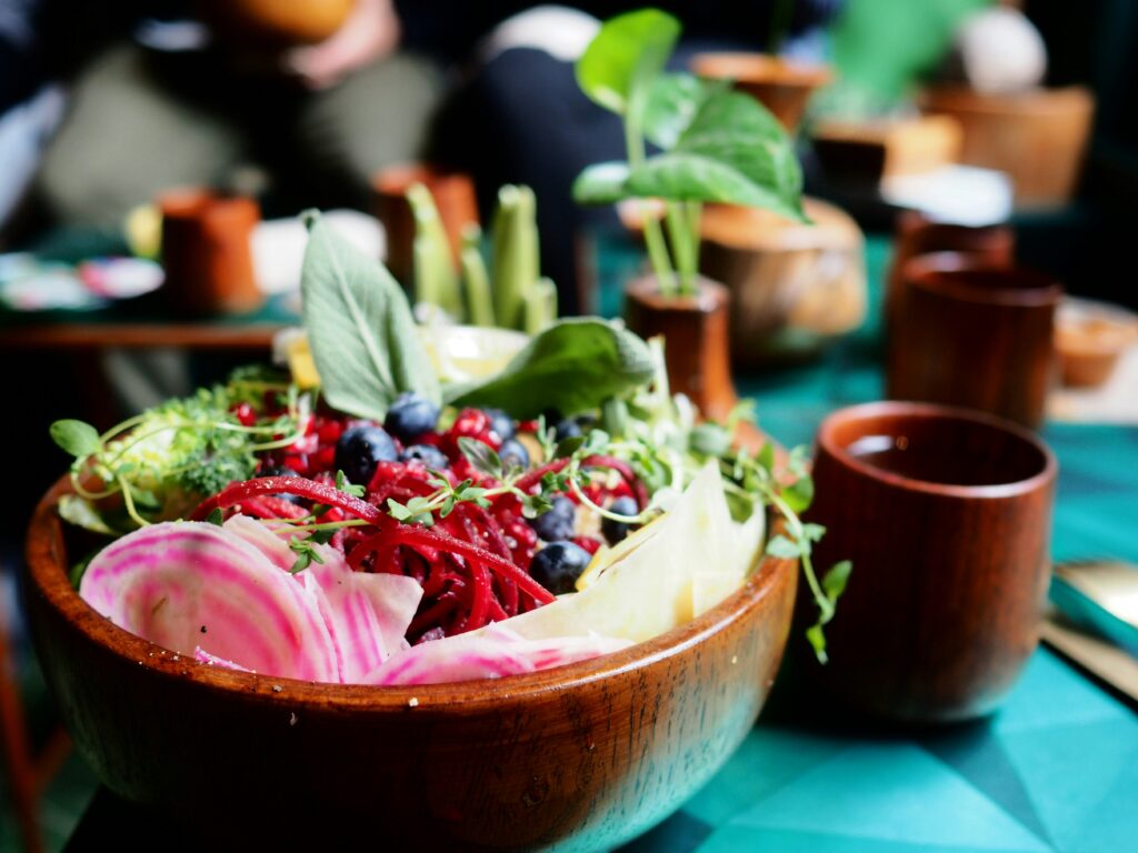 Bunte Salatschüssel aus Holz mit frischem Gemüse und Beeren auf einem gedeckten Tisch. Zu sehen sind unter anderem pink-weiß gestreifte Bete, rote Gemüsestreifen, Blaubeeren, Blattsalat und Sprossen. Im Hintergrund stehen weitere Holzbecher und kleine Pflanzgefäße, die durch die geringe Tiefenschärfe unscharf erscheinen. Die Szene wirkt natürlich, frisch und einladend. | Media Monday #796