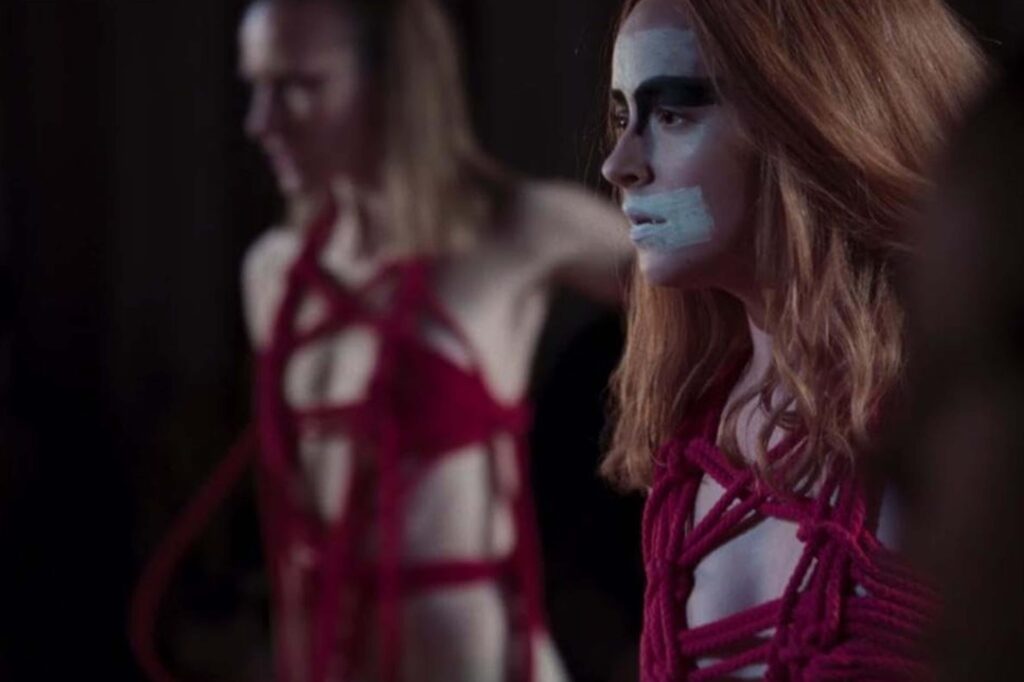 Dakota Johnson (rechts) in Suspiria (2018): Nahaufnahme während einer Tanzszene in einem dunklen Raum. Das Gesicht ist mit weißen Farbstreifen bemalt, während ein rotes Seilkostüm die Figur umhüllt. Eine weitere Tänzerin erscheint unscharf im Vordergrund und verstärkt die beklemmende Atmosphäre der Szene. | Themenübersicht Oktober 2026