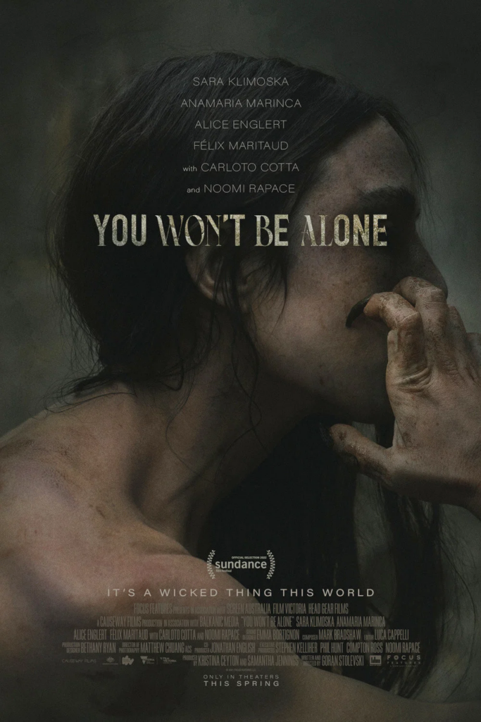 Filmplakat zu You Won’t Be Alone in dunklen, entsättigten Farbtönen. Im Zentrum ist das seitliche Porträt einer Person mit langen, dunklen Haaren zu sehen. Eine Hand liegt vor dem Mund und verdeckt einen Teil des Gesichts. Haut und Hand weisen sichtbare Spuren, Flecken und eine raue Struktur auf. Der Filmtitel steht in großen, hellen Serifenschrift-Buchstaben über dem Gesicht. Oberhalb des Titels sind die Namen der Hauptdarstellenden aufgeführt. Im unteren Bereich befinden sich das Sundance-Festival-Logo, der Werbeslogan „It’s a Wicked Thing This World“ sowie die Produktions- und Besetzungsangaben. Die gedämpfte Farbpalette und die schattenreiche Beleuchtung erzeugen eine düstere, atmosphärische Wirkung.