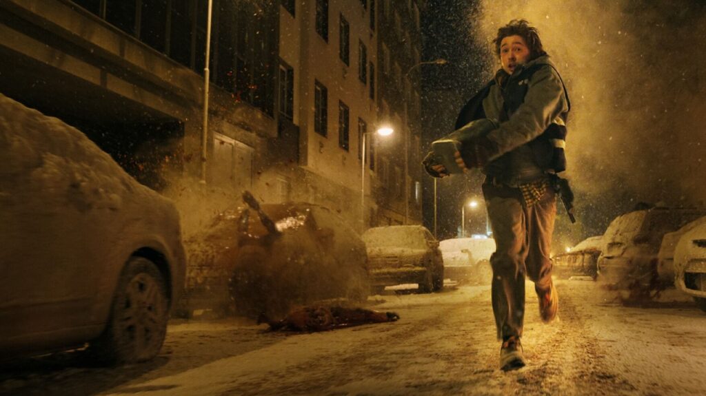 Das Bild zeigt Bryan (Austin Abrams) aus dem Film RESIDENT EVIL, der einen Gegenstand mit beiden Händen trägt und eine beschneite Straße entlang einer Häuserfront und parkenden Autos rennt. Auf der linken Seite des Bildes ist eine Lebensform, die nicht erkennbar ist, gegen ein Auto gekracht und das Blut spritzt und verteilt sich auf Heckscheibe und Motorhaube