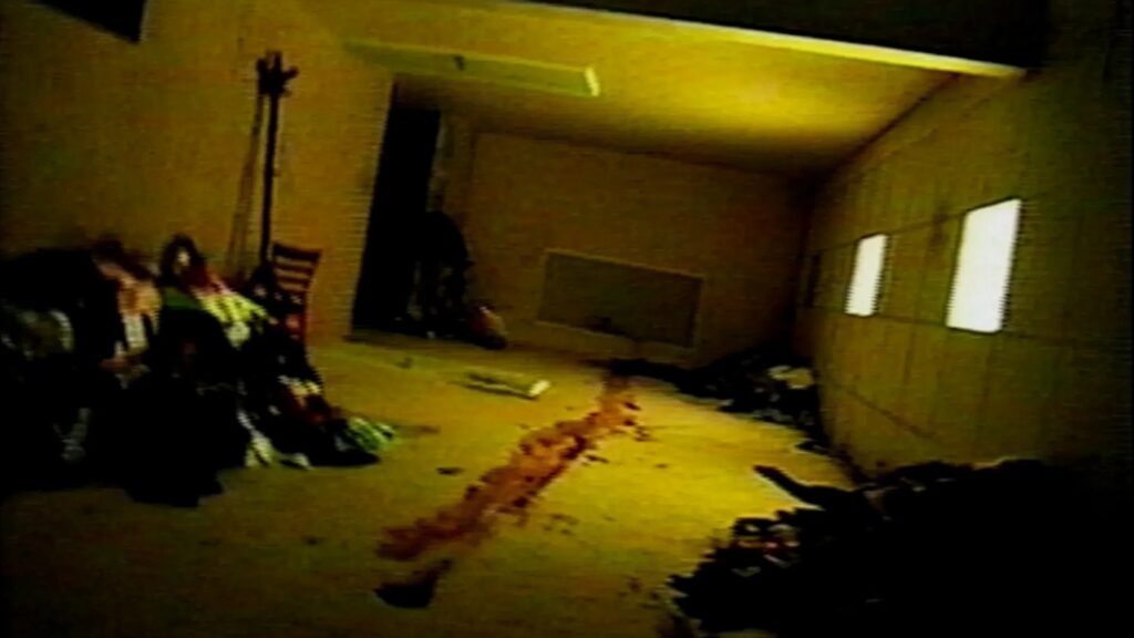 Auf dem Bild ist ein gelber Raum mit Blut auf dem Boden und aufgehäufter Kleiderwäsche zu sehen.
