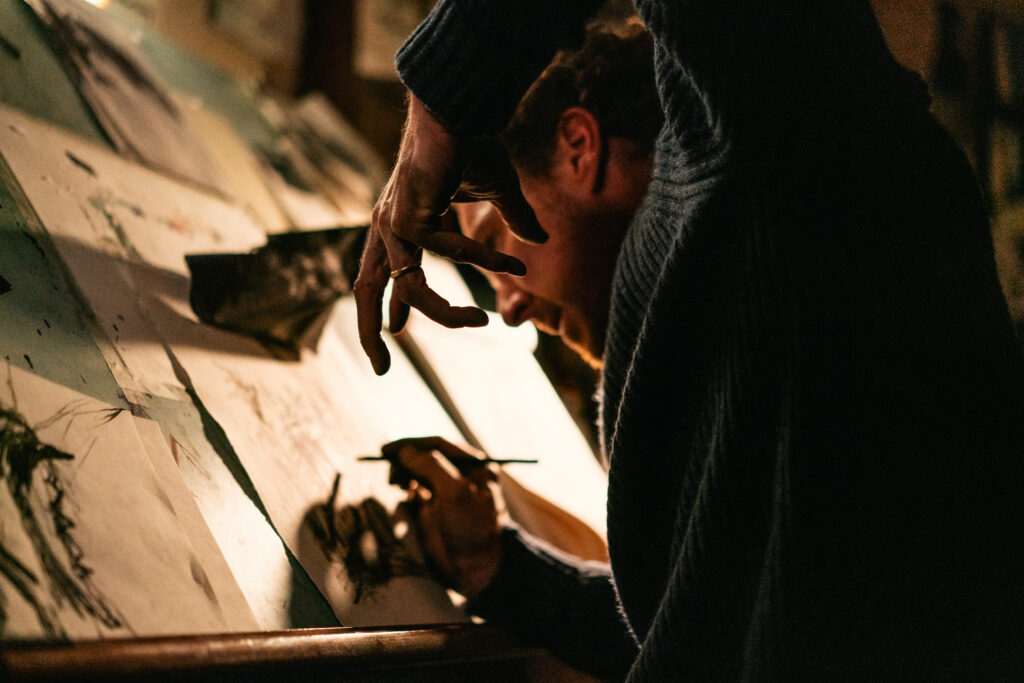Das Bild zeigt Benedict Cumberbatch in "Das Ding mit Federn". Er zeichnet gerade an seinem Zeichentisch und hält die linke Hand wie eine Kralle vor sich