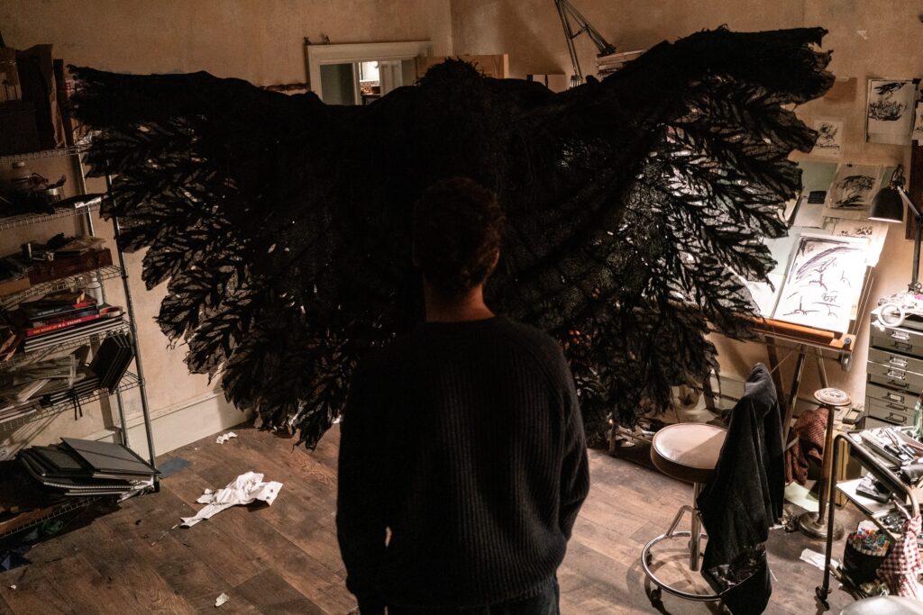 Benedict Cumberbatch mit dem Rücken zu uns, steht vor einem Geschöpf, das aussieht wie eine Krähe. Schwarze ausgebreitete Flügeln wirken als ob sie ihn in eine Umarmung schließen wollen. Die beiden Figuren stehen in einem Atelier | Das Ding mit Federn