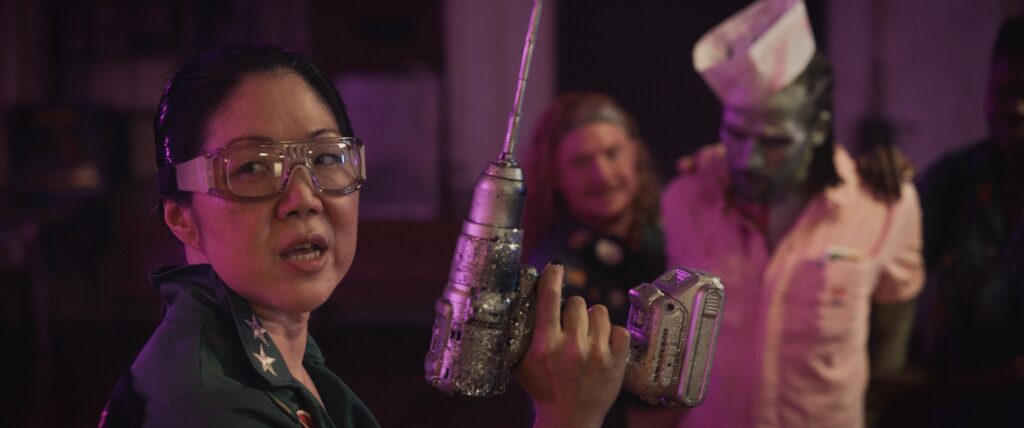 Eine Person mit einem Bohrer schaut in die Kamera. Im Hintergrund halten zwei weitere Personen einen Zombie fest. | Queens of the Dead – Filmkritik