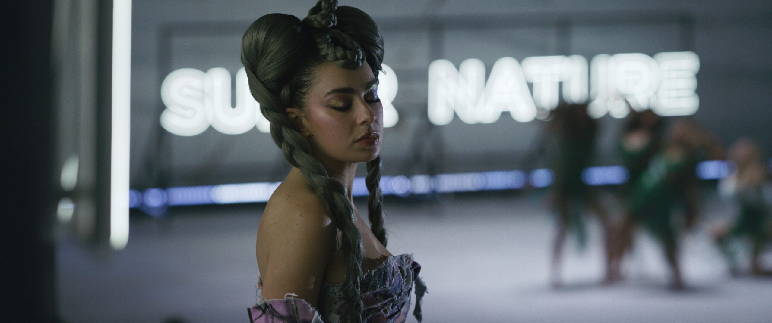 Das Bild zeigt Charli xcx (Mother Nature) mit geschlossenen Augen vor einem Neonschriftzug stehen, der im Hintergrund verschwimmt