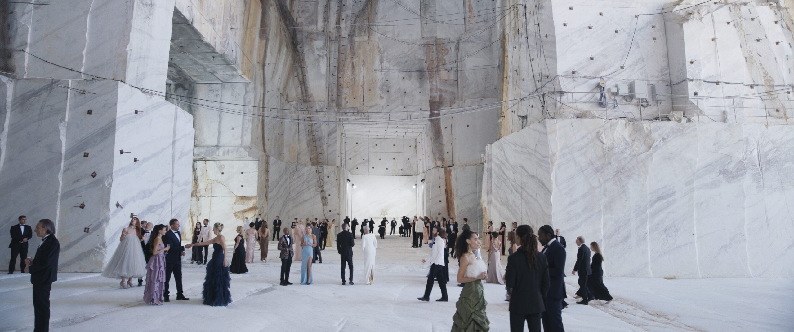 Elegante Gesellschaft aus dem Film SACRIFICE in Abendgarderobe versammelt sich in einer riesigen, hellen Marmorhalle mit hohen, roh wirkenden Steinwänden. Die monumentale Innenarchitektur wird von gewaltigen Marmorblöcken, feinen Seil- und Kabelstrukturen sowie einem zentralen Durchgang geprägt. Die Menschen stehen verteilt im Raum, unterhalten sich oder bewegen sich durch die weitläufige Kulisse, die an einen Steinbruch oder eine unterirdische Kathedrale erinnert.