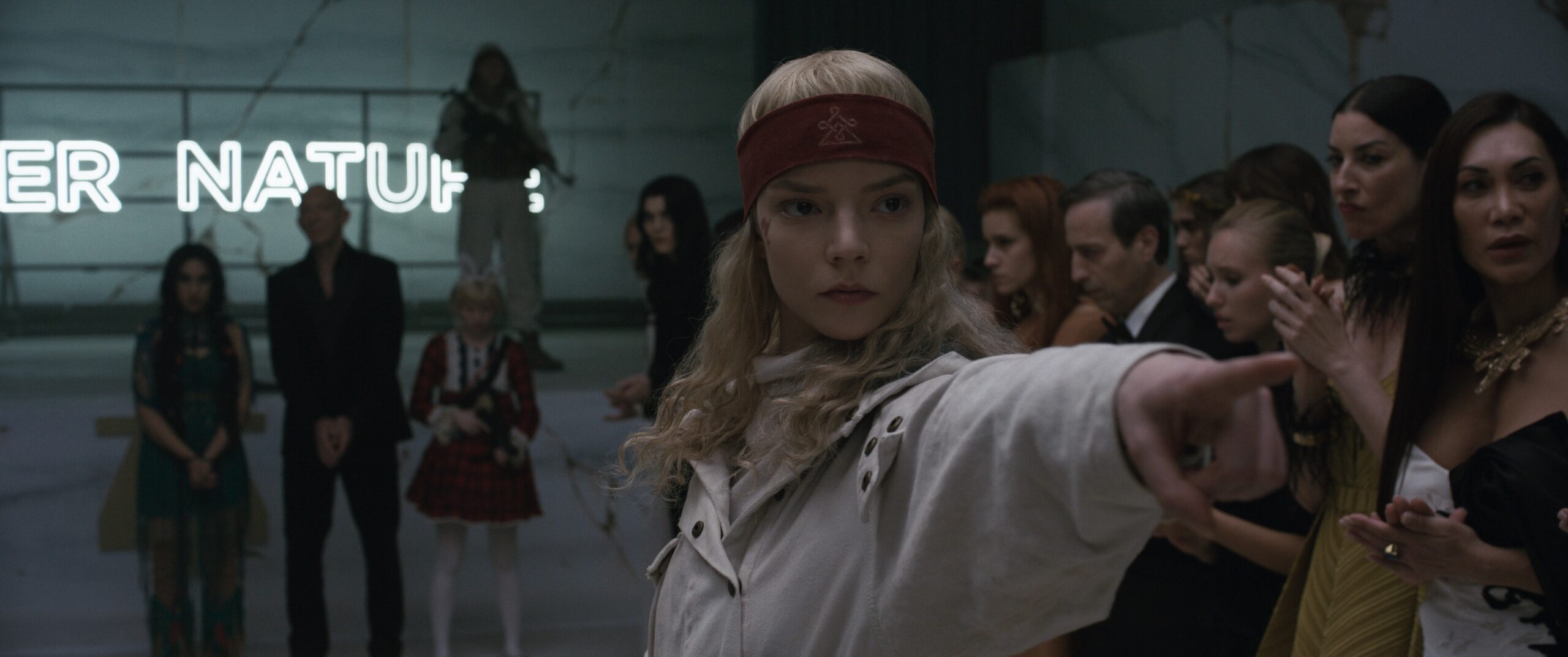 Das Bild zeigt Joan (Anya Taylor-Joy) aus dem Film SACRIFICE vor einer Menschenmenge stehen. Im Hintergrund ist ein Neonschriftzug mit dem Slogan "Natur" zu sehen. Sie zeigt mit dem Finger in eine Richtung außerhalb des Bildes