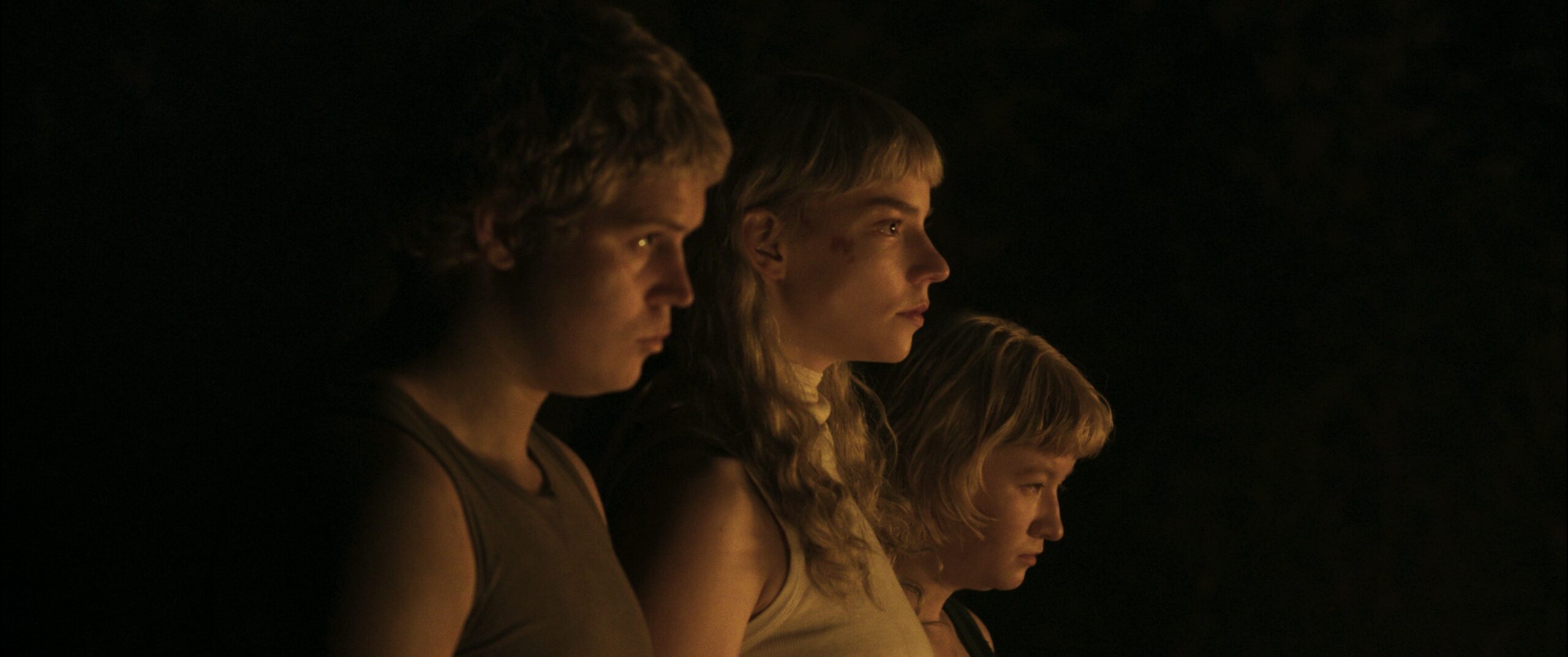 Das Bild zeigt von links: Jonatan „Yung Lean“ Leandoer, Anya Taylor-Joy und Jade Croot nebeneinander im orangebraunen Licht stehen. Der Anblick hat etwas mystisches, sehr passend zum Film SACRIFICE