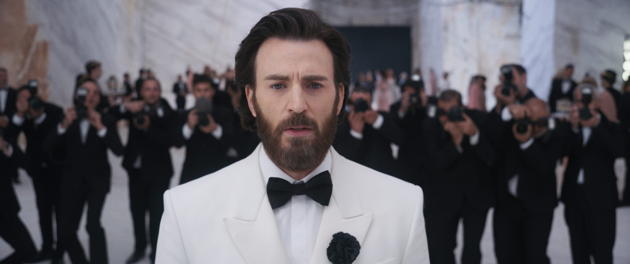 Das Bild zeigt Mike Tyler (Chris Evans) im weißen Jackett vor einer menge mit Fotograf:innen stehen, die alle die Kamera hochhalten und durchgehend schwarze Anzüge tragen. Sein Blick ist zweifelnd und nachdenklich