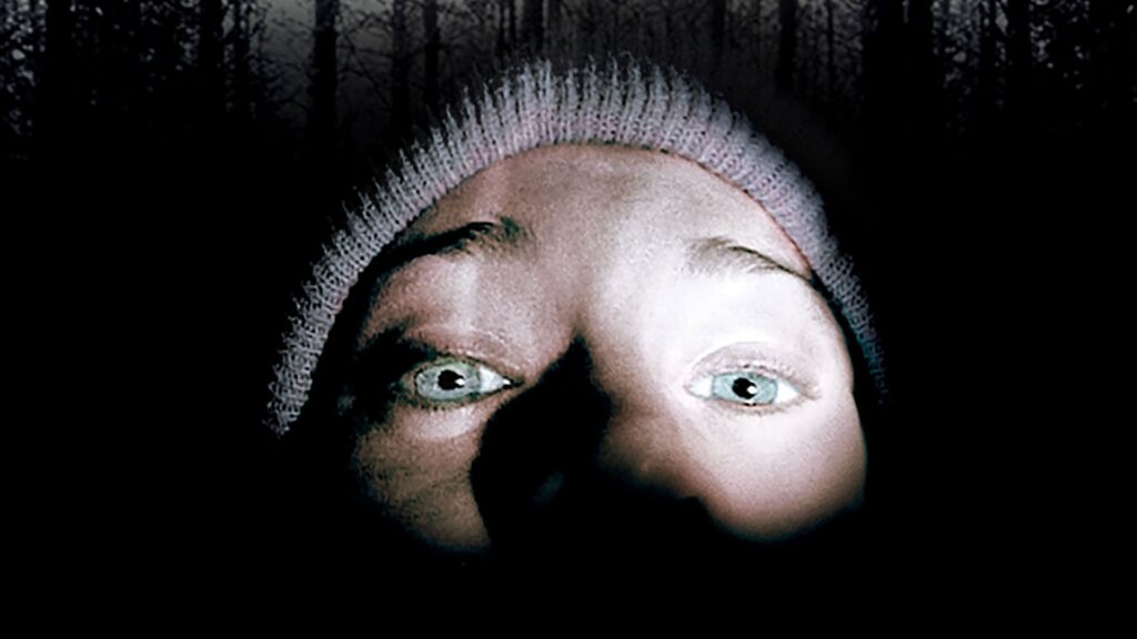 Die obere Gesichtshälfte einer Person ist in Nahaufnahme zu sehen. Im Hintergrund ist Wald angedeutet, ansonsten Schwärze.