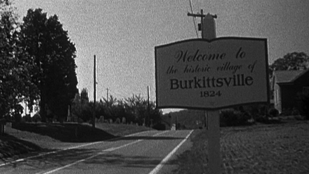 Ein schwarz-weißes Foto mit einer Straße und einem Ortseingangsschild: "Welcome to the historic village of Burkittsville 1824".